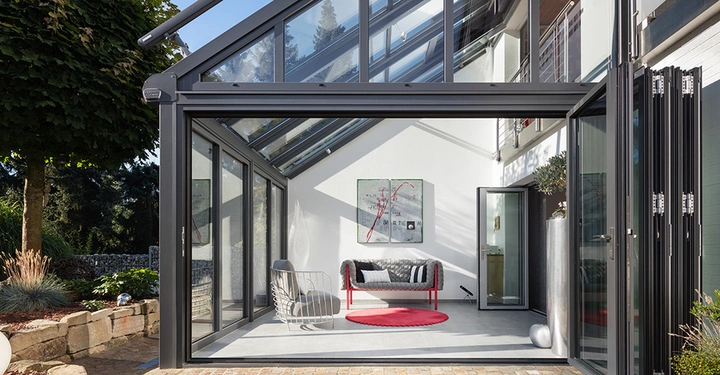 Galerie-Bild 4: Wintergarten mit moderner Möblierung umfasst Sofa und Sessel auf grauem Boden vor weißer Wand darunter zwei Kunstwerke mit abstrahierten roten Linien im Außenbereich eines zeitgenössischen Hauses umgeben von Pflanzen.