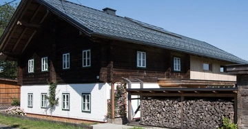 Ein traditionelles Holzhaus steht ruhig in einer sonnigen ländlichen Umgebung mit gestapeltem Feuerholz unter einem überdachten Bereich vor dem Haus und blühenden Pflanzen entlang der Fassade.