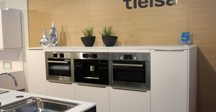 Galerie-Bild 12: Moderne Küchengeräte in glänzendem Schwarz eingebaut in weiße Schränke darüber Blumenkübel und Vasen dahinter Holzwand mit Beschriftung TIELSA und Schild mit Aufschrift 5 JAHRE VOLLGARANTIE