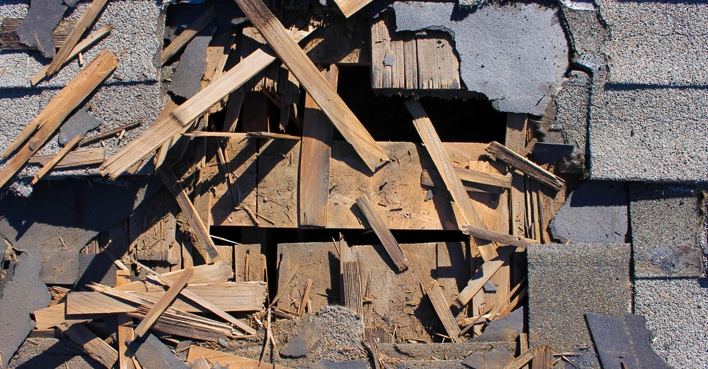 Ein Holz-durchbohrtes Dach zeigt beschädigte Schindeln und Holzlatten, umgeben von zerschlagenem Baumaterial, auf einer grauen, kiesbedeckten Oberfläche.