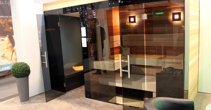 Galerie-Bild 21: Saunakabine in modernem Design steht geöffnet in einem Ausstellungsraum mit Teppichboden. Innenausstattung aus Holz mit Handtüchern. Text auf der Kabine home spa S.