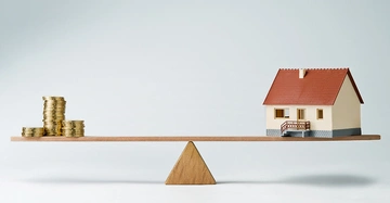 Haus und Münzstapel stehen auf einem hölzernen Schaukelbrett balancierend auf einem dreieckigen Holzblock in minimalistischer Umgebung