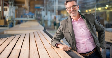 Mann lehnt an einem Stapel Holzlatten in einer großen Werkstatt Fabrikhalle mit industriellen Maschinen und Holzmaterialien im Hintergrund trägt eine Brille ein rosa Hemd und ein graues Sakko