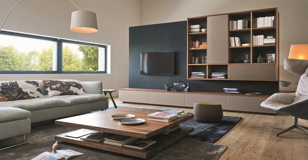 Sofa mit bunten Kissen steht vor einem großen Fenster neben einem niedrigen Couchtisch auf Teppich in modernem Wohnzimmer mit Wandregalen Fernseher und Stehlampe auf Holzboden.