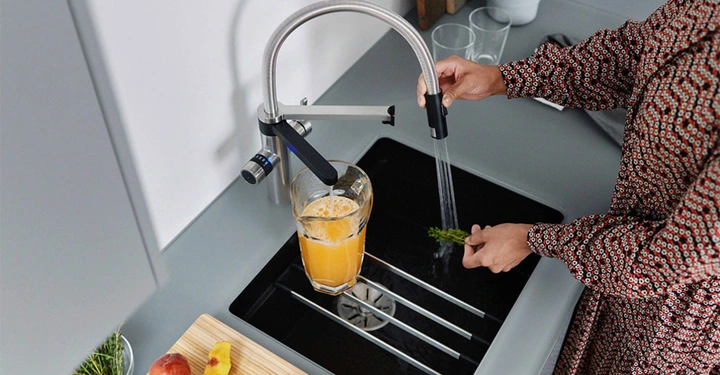 Eine Person wäscht Gemüse unter einem Wasserhahn in einer modernen Küche. Im Waschbecken steht eine Saftkanne. Auf dem Schneidebrett liegen eine Pfirsichhälfte und geschnittene Stücke.