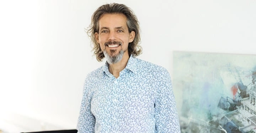 Ein Mann mit langen Haaren und Bart lächelt in die Kamera Er trägt ein hellblau-weißes gemustertes Hemd Im Hintergrund ist ein Kunstwerk an einer weißen Wand zu sehen