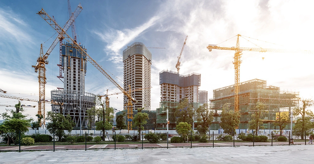 Baukräne errichten Hochhäuser in einem städtischen Gebiet umgeben von Baugerüsten und Bäumen bei sonnigem Himmel.