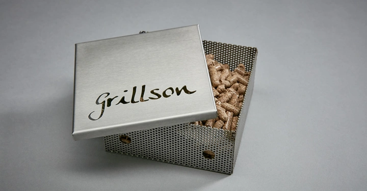 Metallbox mit Gravur Grillson auf dem Deckel teilweise geöffnet gefüllt mit Holzpellets auf grauem Hintergrund
