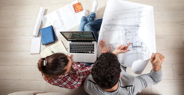 Pärchen von oben fotografiert sitzt am Boden, beugt sich über Wohnungspläne, Laptop am Schoß. Wohnung leer,