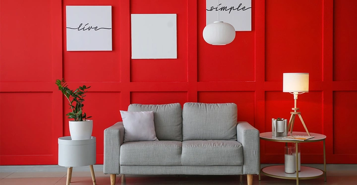 Hellgraue Zweisitzercouch vor knallroter Wand, auf der drei weiße Bilder hängen. Neben dem Sofa rechts steht eine Stehleuchte, links eine große Topfpflanze.