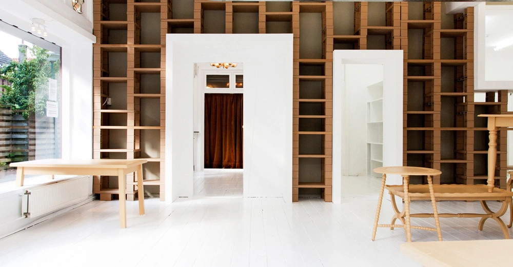 Leerer Raum mit Bücherregalen an der Wand Holztisch und Bank im Vordergrund durch große Fenster flutet Tageslicht herein Holzregale und weiß gestrichener Boden schaffen eine helle Atmosphäre.