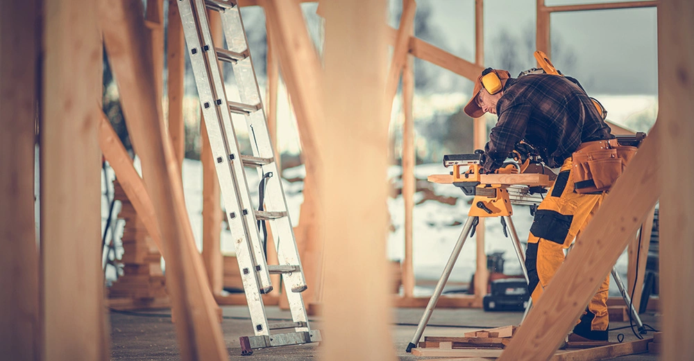 Arbeiter schneidet im Rohbau eines Holzhauses auf einer mobilen Werkbank ein Stück Holz zu. Er trägt ein dunkles, kariertes Hemd, einen gelben Gehörschutz, eine gelbe Kappe und eine gelbe Arbeitshose.