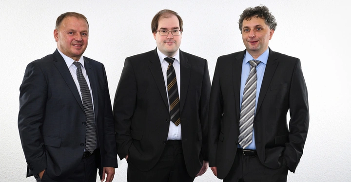 Drei Männer stehen in Anzügen nebeneinander in einem weißen Raum. Der linke Mann lächelt, der mittlere trägt eine Brille, der rechte Mann hat lockiges Haar.