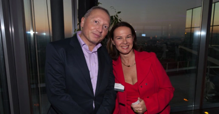 Galerie-Bild 53: Zwei Personen stehen nebeneinander und lächeln kamera. Sie befinden sich in einem modernen Innenraum mit großen Fenstern im Hintergrund die eine Aussicht auf eine Stadtlandschaft bei Dämmerung zeigen.