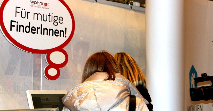 Galerie-Bild 9: Zwei Personen stehen an einem Stand und betrachten ein Infoterminal im Innenbereich eines Messegeländes. Ein Schild darüber zeigt den Text wohnnet Für mutige FinderInnen.