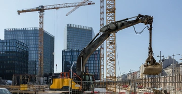 Baumaschine hebt Last auf Baustelle mit Hochhäusern im Hintergrund Kräne ragen in den Himmel daneben.