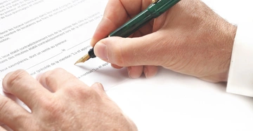 Hand hält grünen Stift unterschreibt Dokument Text auf Dokument enthält Absätze und leere Zeilen um Datum und Unterschrift zu platzieren die Umgebung ist weiß und hell.