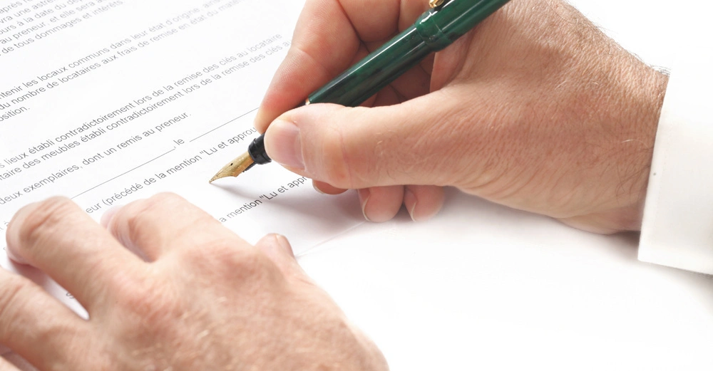 Hand hält grünen Stift unterschreibt Dokument Text auf Dokument enthält Absätze und leere Zeilen um Datum und Unterschrift zu platzieren die Umgebung ist weiß und hell.