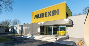 Gebäude mit großem gelben Schild MUREXIN darüber graues Gebäude Technikum auf der Fassade treppenführend zu einem gläsernen Eingang umgeben von einer gepflasterten Einfahrt und grasbewachsenen Außenflächen Bäume im Hintergrund.