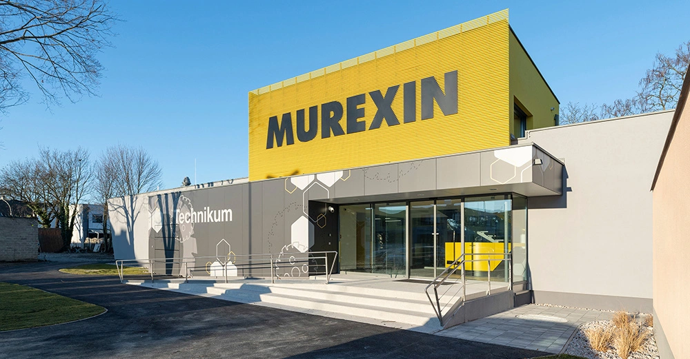 Gebäude mit großem gelben Schild MUREXIN darüber graues Gebäude Technikum auf der Fassade treppenführend zu einem gläsernen Eingang umgeben von einer gepflasterten Einfahrt und grasbewachsenen Außenflächen Bäume im Hintergrund.