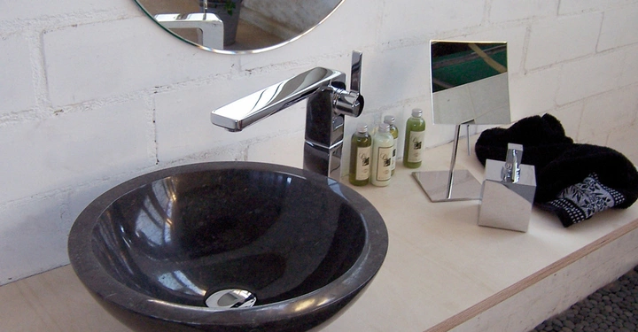 Galerie-Bild 4: Dunkles Rundwaschbecken mit moderner Armatur, umgeben von Toilettenartikeln und Spiegeln, auf einer hellen Holzplatte vor einer weißen Backsteinmauer. Im Hintergrund steht eine Pflanze.