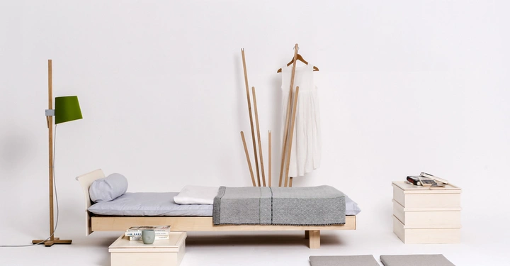 Galerie-Bild 4: Holzbett mit grauer Bettwäsche steht in einem minimalistisch eingerichteten, weißen Raum; daneben eine Stehlampe, zwei Polster auf dem Boden und zwei Stapel Holzkisten mit Büchern.