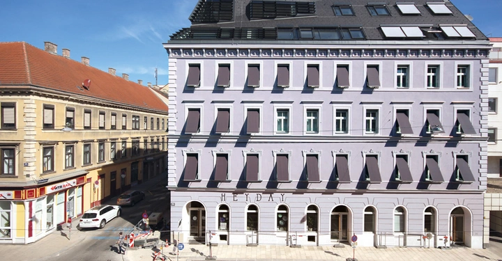 Galerie-Bild 14: Gebäude mit lilafarbener Fassade und vielen Fenstern Sonnenschirme an den Fenstern Straße mit Gehweg und Autos im Vordergrund andere Gebäude im Hintergrund Text: HEYDAY
