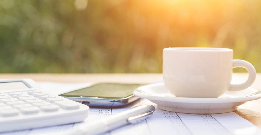 Weiße Kaffeetasse steht auf einem Unterteller auf Dokumenten neben einem Taschenrechner und einem Stift in einer sonnigen Outdoor-Umgebung mit unscharfem grünem Hintergrund