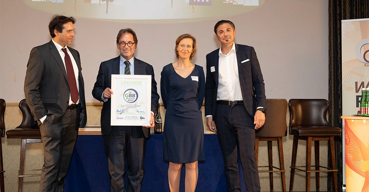 Galerie-Bild 9: Vier Menschen posieren mit einer Urkunde in einem formellen Umfeld. Die Urkunde trägt den Titel URKUNDE GBB Award 2021 und zeigt Unterschriften. Im Hintergrund sind Stühle und eine Leinwand.