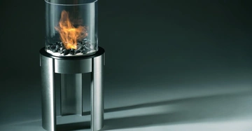 Metallischer Standfuß mit Glaszylinder enthält Feuer umgeben von schwarzen Steinen auf grauem Hintergrund