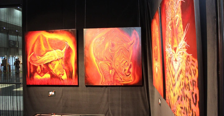 Galerie-Bild 16: Bunte Malereien von Tieren hängen an einer schwarzen Wand in einer Kunstgalerie. Ein Tisch mit Glasplatte steht davor. Zwei Personen sind im Hintergrund sichtbar.