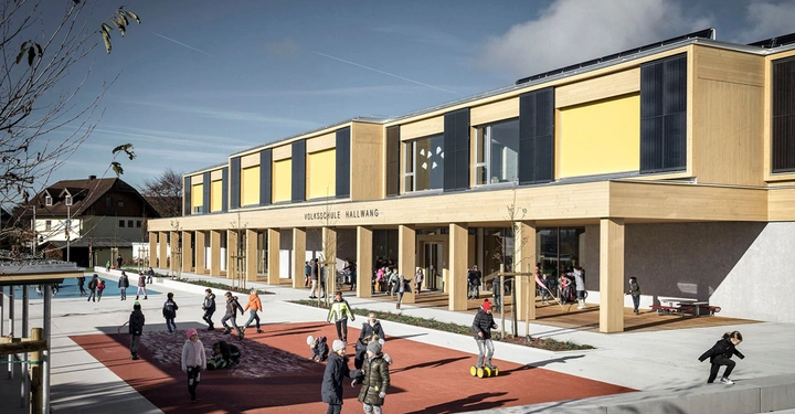 Galerie-Bild 10: Mehrere spielende Kinder rennen und spielen vor einem zweistöckigen modernen Schulgebäude aus Holz auf einem Spielplatz an einem sonnigen Tag VOLKSSCHULE HALLWANG ist an der Fassade zu lesen