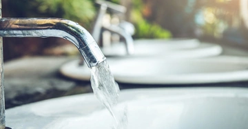 Ein Wasserhahn lässt Wasser in ein Waschbecken fließen während im Hintergrund weitere Waschbecken und Pflanzen zu sehen sind
