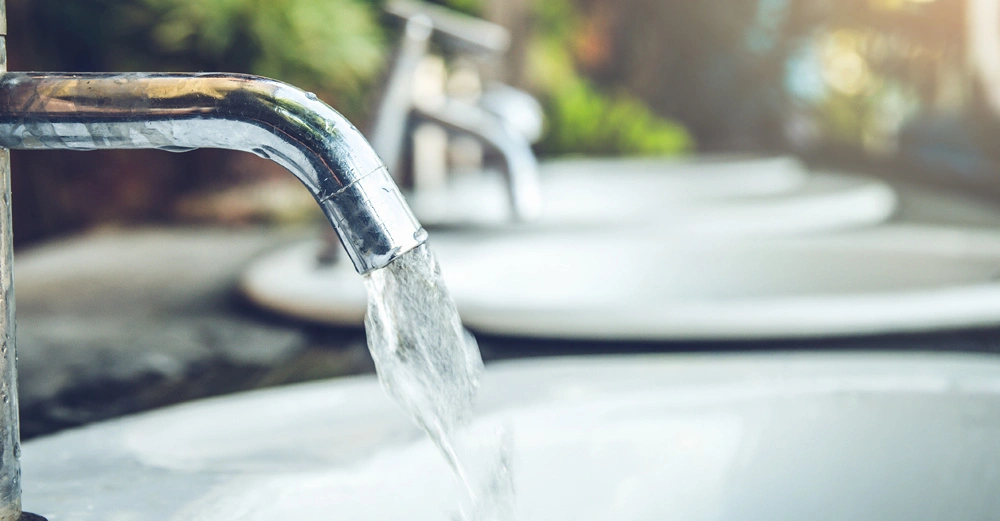 Ein Wasserhahn lässt Wasser in ein Waschbecken fließen während im Hintergrund weitere Waschbecken und Pflanzen zu sehen sind