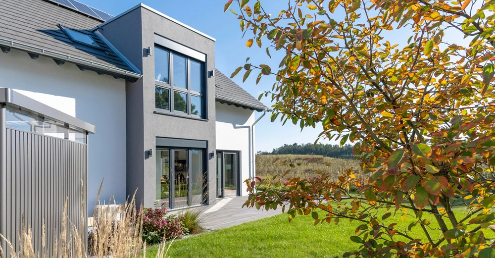Modernes Einfamilienhaus mit dunkelgrau gestrichenem Erker im Grünen. Rechts im Bild ein herbstlich verfärbter Laubbaum, links ein Gartenhäuschen aus Alu.
