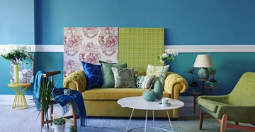 Gelbes Sofa mit bunten Kissen steht vor blauen Wänden mit weißen Leisten und farbigem Wandkunstwerke dekoriert Ein weißer Tisch und grüne Sessel umgeben von Zimmerpflanzen und einer Lampe vervollständigen das Wohnzimmer.