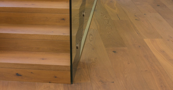 Holztreppe mit Glasgeländer steigt nach oben in einem modernen Innenraum mit hellbraunem Holzboden.