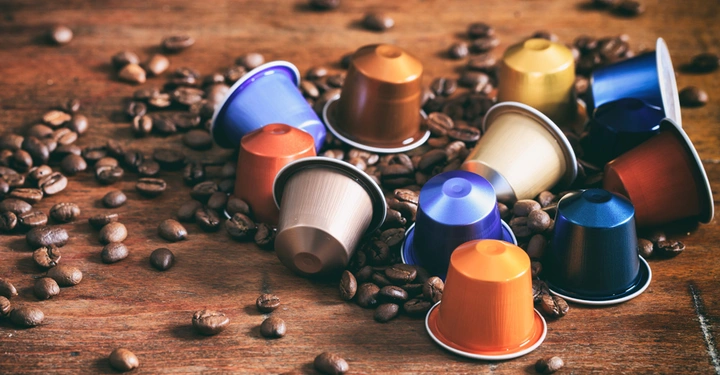 Mehrfarbige Kaffeekapseln liegen verstreut auf einem hölzernen Tisch zwischen zahlreichen Kaffeebohnen.