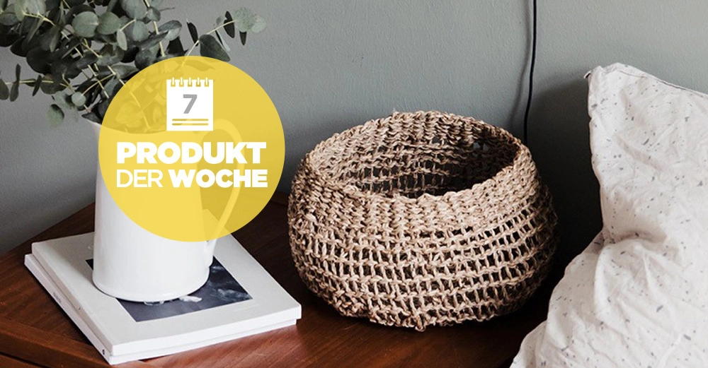 Ein Korb steht neben einem Kissen auf einem Holztisch Daneben befindet sich eine weiße Kanne mit Zweigen und Büchern Text 7 PRODUKT DER WOCHE