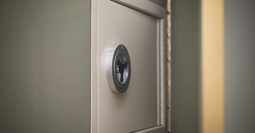 Ein kleiner Tresor mit Drehknopf ist an einer grünen Wand montiert. Der Tresor ist leicht geöffnet, wobei das Scharnier und ein Teil des Inneren sichtbar sind.