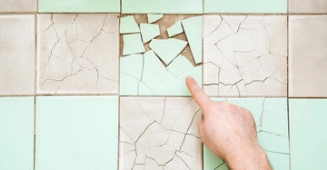 Hand zeigt auf eine türkise zerbrochene Fliese in einem türkis-beigen Badezimmerboden.
