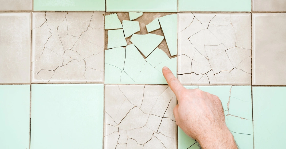 Hand zeigt auf eine türkise zerbrochene Fliese in einem türkis-beigen Badezimmerboden.