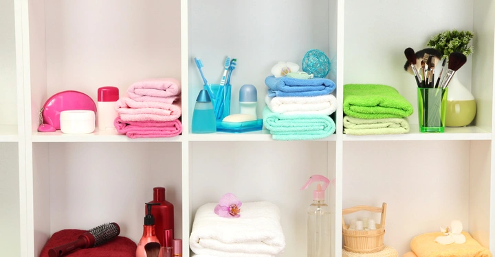 Zusammengefaltete Handtücher in verschiedenen Farben liegen auf weißen Regalen; daneben sind Kosmetikartikel wie Cremes, Zahnbürsten, Parfums und Makeup-Pinsel angeordnet; die Regale befinden sich in einem Badezimmer.