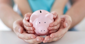 Ein rosa Sparschwein wird von kleinen Kinderhänden gehalten, die wiederum von größeren Erwachsenen händen gestützt werden, auf einem weißen Tisch in einem hellen Raum