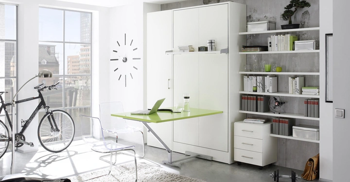 Ein Schreibtisch mit offenem Laptop steht in einem hellen Raum neben großen Fenstern links ein Fahrrad sowie transparente Stühle rechts Bücherregale mit Büchern und Dekoartikeln daneben ein Schrank.