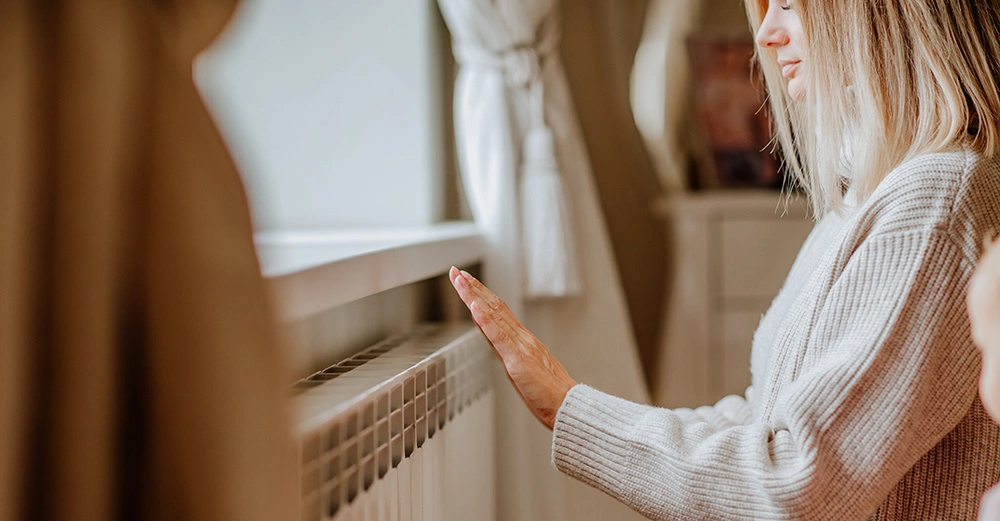 blonde Frau mit weißem Wollpullover hält ihre Hand an warmen Heizkörper