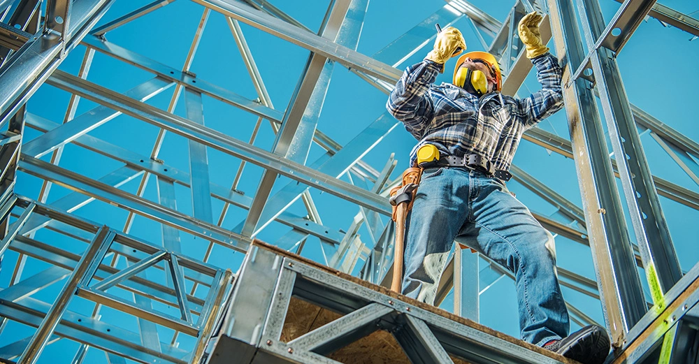 Bauarbeiter trägt Schutzhelmausrüstung, hebt Werkzeuge mit beiden Händen hoch, arbeitet an einer Baustelle aus Metallträgern unter blauem Himmel.