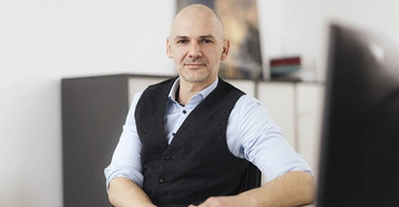 Ein Mann sitzt lächelnd am Schreibtisch in einem Büro trägt ein blaues Hemd und eine dunkle Weste im Hintergrund befinden sich Regale mit Büchern und Ordnern