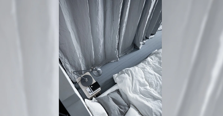 Galerie-Bild 7: Bett mit zerwühlter weißer Bettwäsche und grauen Kissen steht neben einem Nachttisch mit einer Lampe und Büchern Vorhänge mit grobem Stoff hängen im Hintergrund.