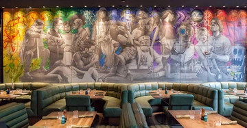 Wandgemälde zeigt Gruppen von Menschen in verschiedensten Szenen und Stilen an einer Wand eines Restaurants mit grünen, gepolsterten Sitzbereichen und gedeckten Holztischen im Vordergrund.